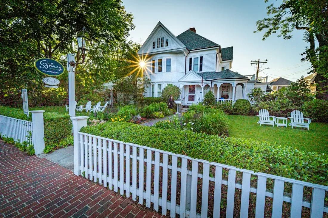 White picket fence along a brick path in front of a large house and garden.