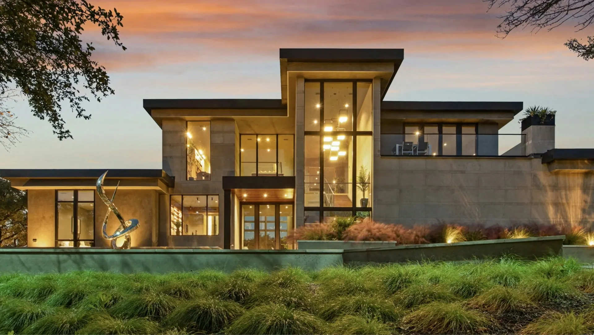 Modern two-storey home at dusk with tall lit windows, stone and timber cladding, and ornamental grasses in front.