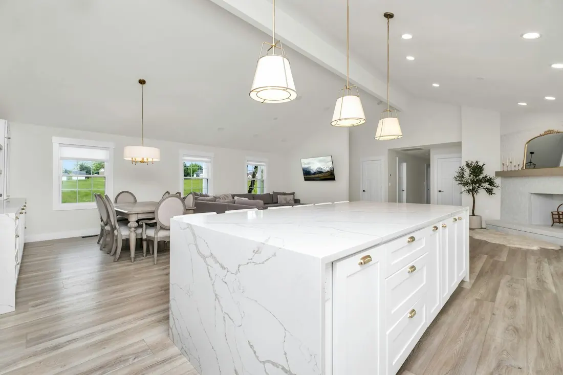 Open-plan kitchen with a large marble-topped island, white cabinets, pendant lights and a living area beyond.