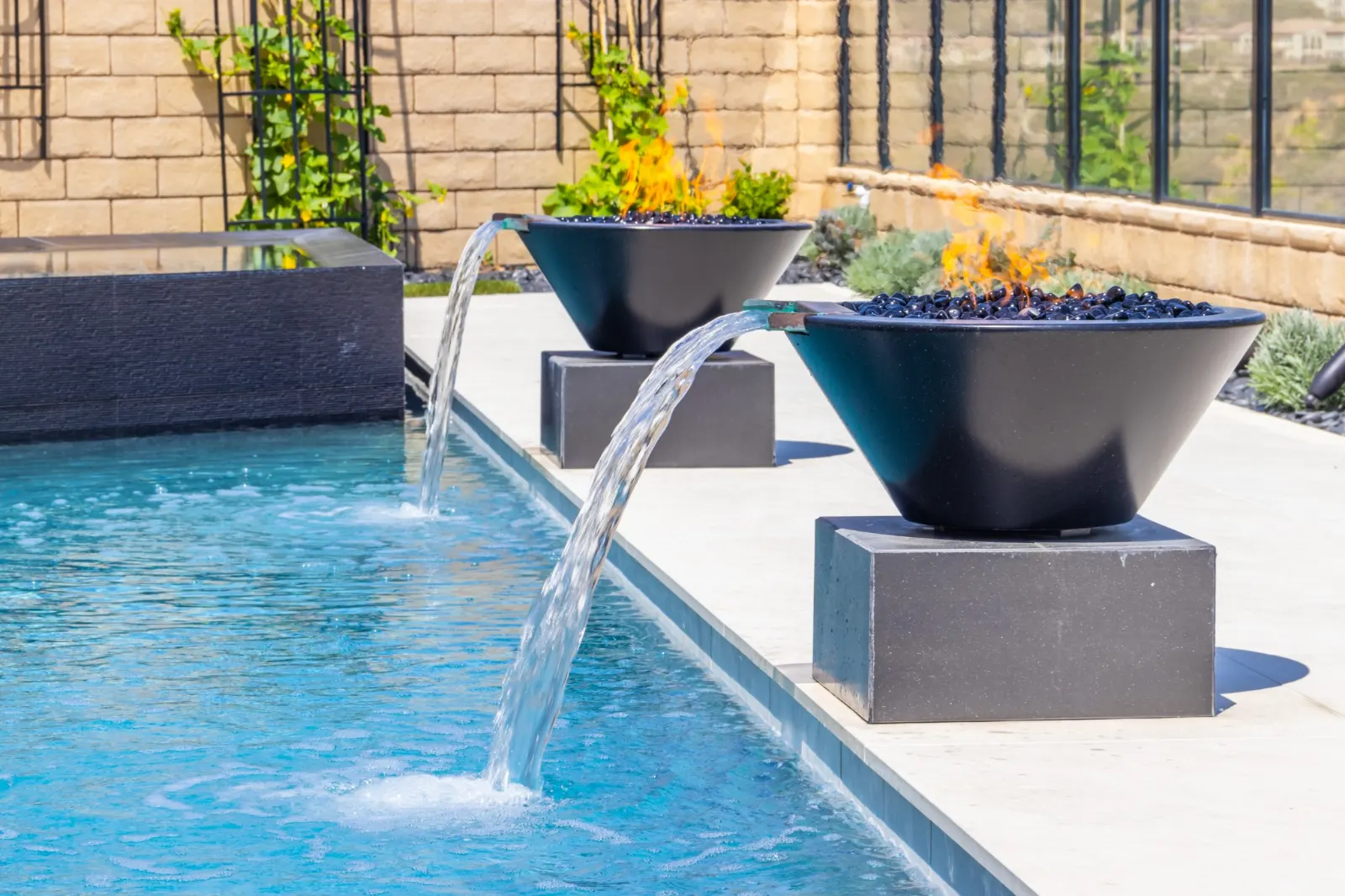 Two dark bowls set on a pool wall, each spilling a sheet of water into the pool below.