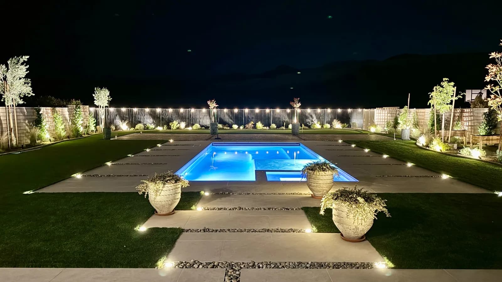 Swimming pool lit at night, with illuminated planters and lighting along a symmetrical deck.