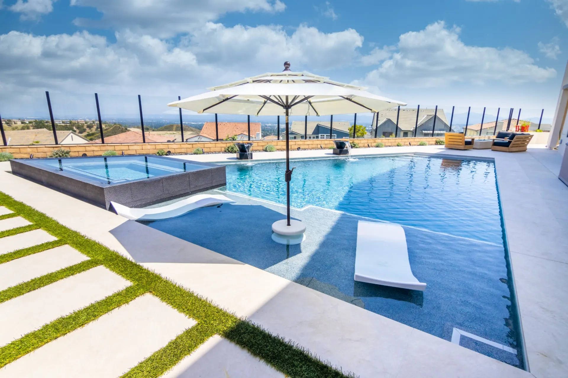 Rectangular swimming pool with a raised spa, a tanning shelf, a large patio umbrella and lounge chairs on a paved deck.