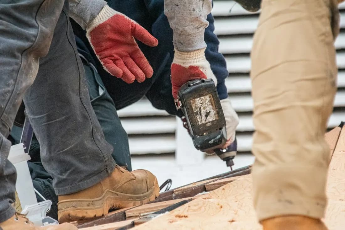 Two workers in gloves and work boots fastening material on a roof with a power drill.
