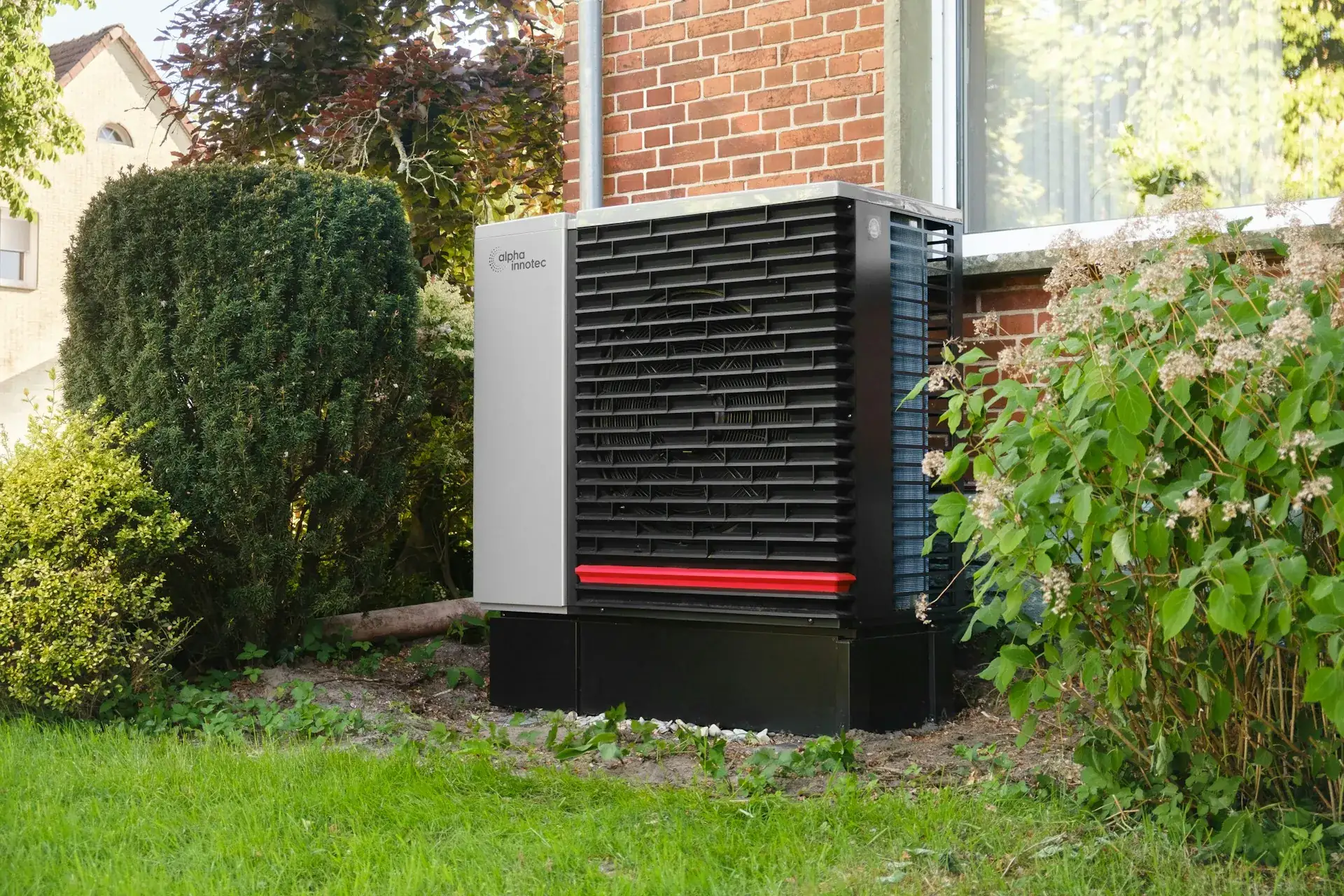 Modern outdoor heat pump unit on a gravel bed beside a brick house, framed by shrubs.