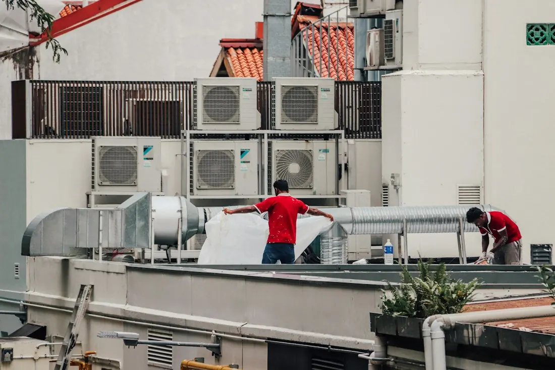 Two workers beside a bank of outdoor air-conditioning condenser units and ducting.