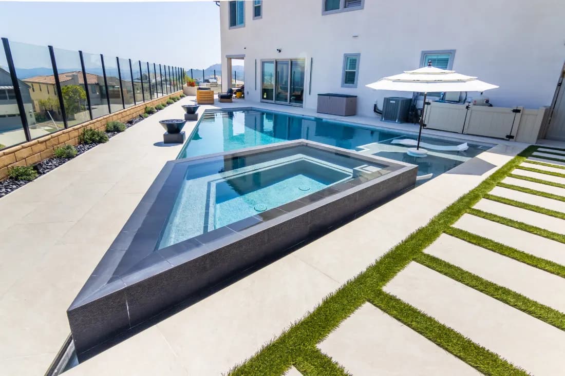 Long swimming pool with an angled spa, tiled edges, turf joints between paving and a patio umbrella.