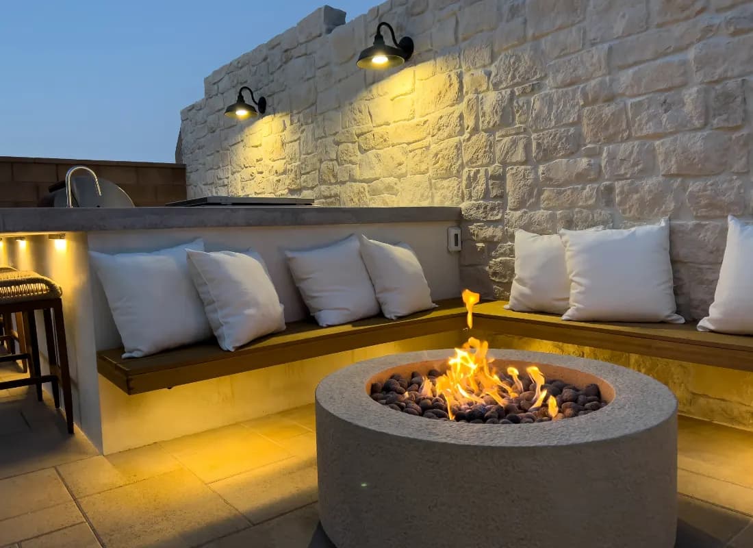 Round gas fire pit with cushioned bench seating against a stone wall on a wood deck.